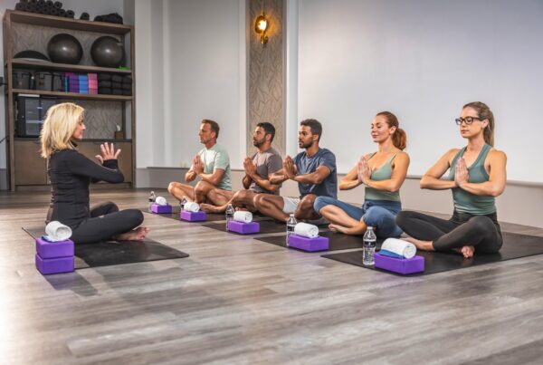 A yoga instructor leads five adults seated on mats in a studio, all in a cross-legged position with hands in prayer pose, facing the instructor. Yoga props and water bottles are placed beside them.
