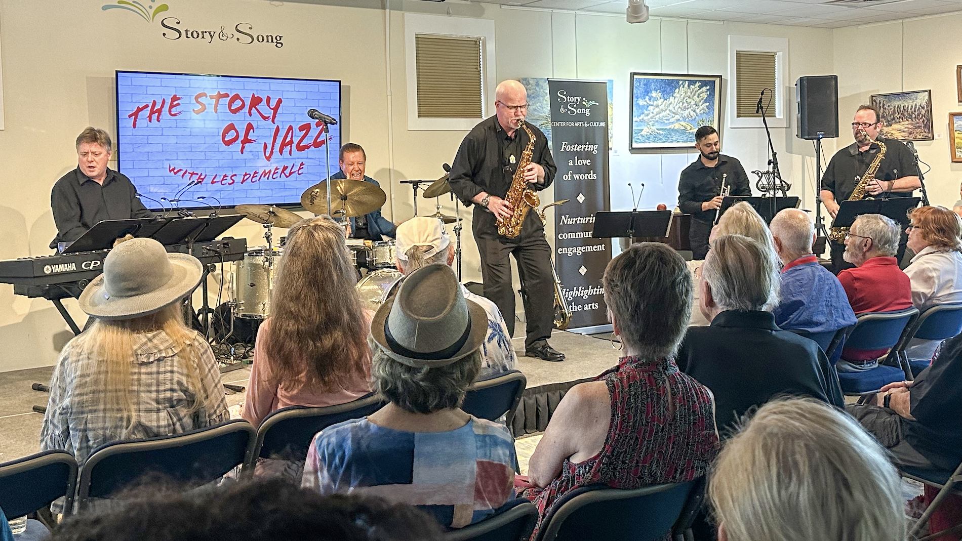 A jazz band performs on stage for an audience, with a screen behind them displaying The Story of Jazz with Les DeMerle. The audience sits in rows, attentively watching the musicians play.