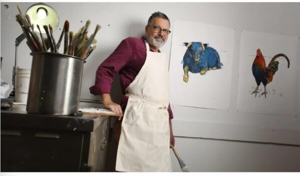 A man with gray hair and glasses, wearing a white apron over a maroon shirt, stands in an art studio beside a table of paintbrushes. Animal paintings of a blue cow and a rooster hang on the wall behind him.