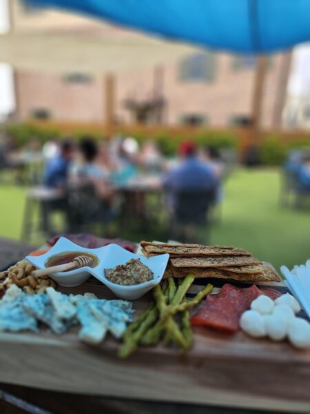 A wooden platter with cheese, crackers, nuts, pickles, spread, and cured meats in focus, with a blurred outdoor scene of people sitting at tables in the background.