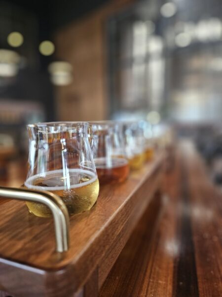 A close-up view of a wooden tray holding several small glasses filled with different types of beer, arranged in a row, inside a softly lit bar or brewery.
