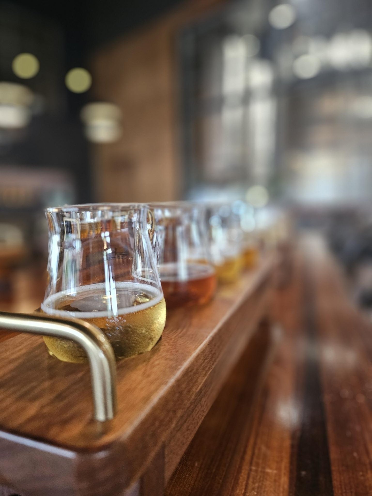 A close-up view of a wooden tray holding several small glasses filled with different types of beer, arranged in a row, inside a softly lit bar or brewery.