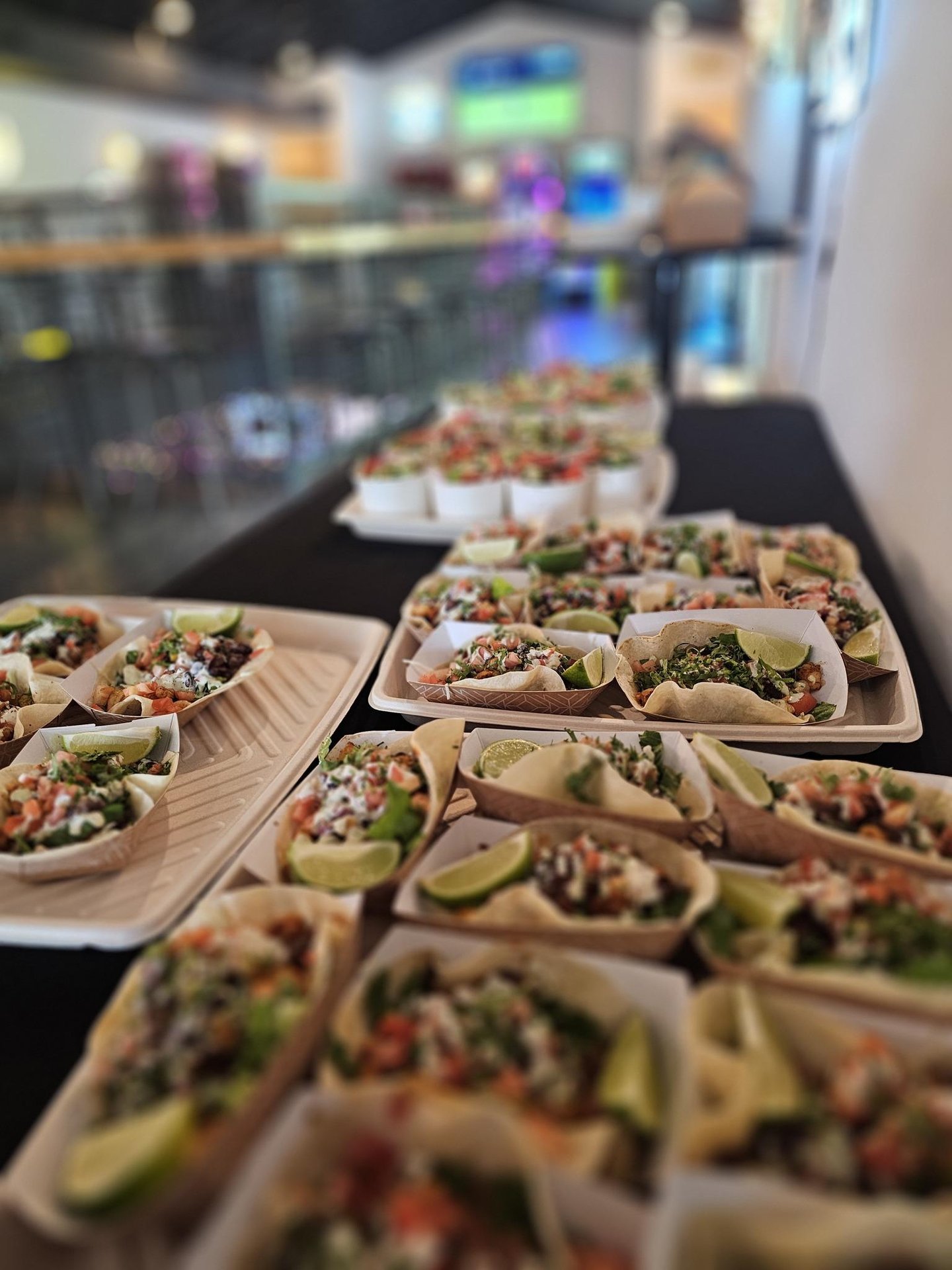 A close-up of trays and plates filled with assorted tacos topped with fresh vegetables, lime wedges, and various toppings, displayed on a black tablecloth in a brightly lit indoor setting.