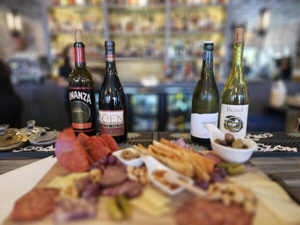 A charcuterie board with assorted meats, cheeses, nuts, crackers, and olives sits in the foreground, with three bottles of wine displayed behind it on a bar counter in a restaurant.