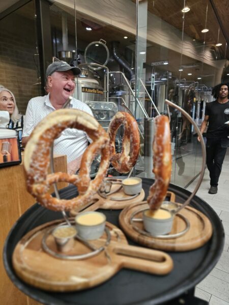 A smiling man stands behind a table holding large pretzels on metal stands, each served with small cups of dipping sauce, in a modern restaurant with glass walls and brewing equipment in the background.