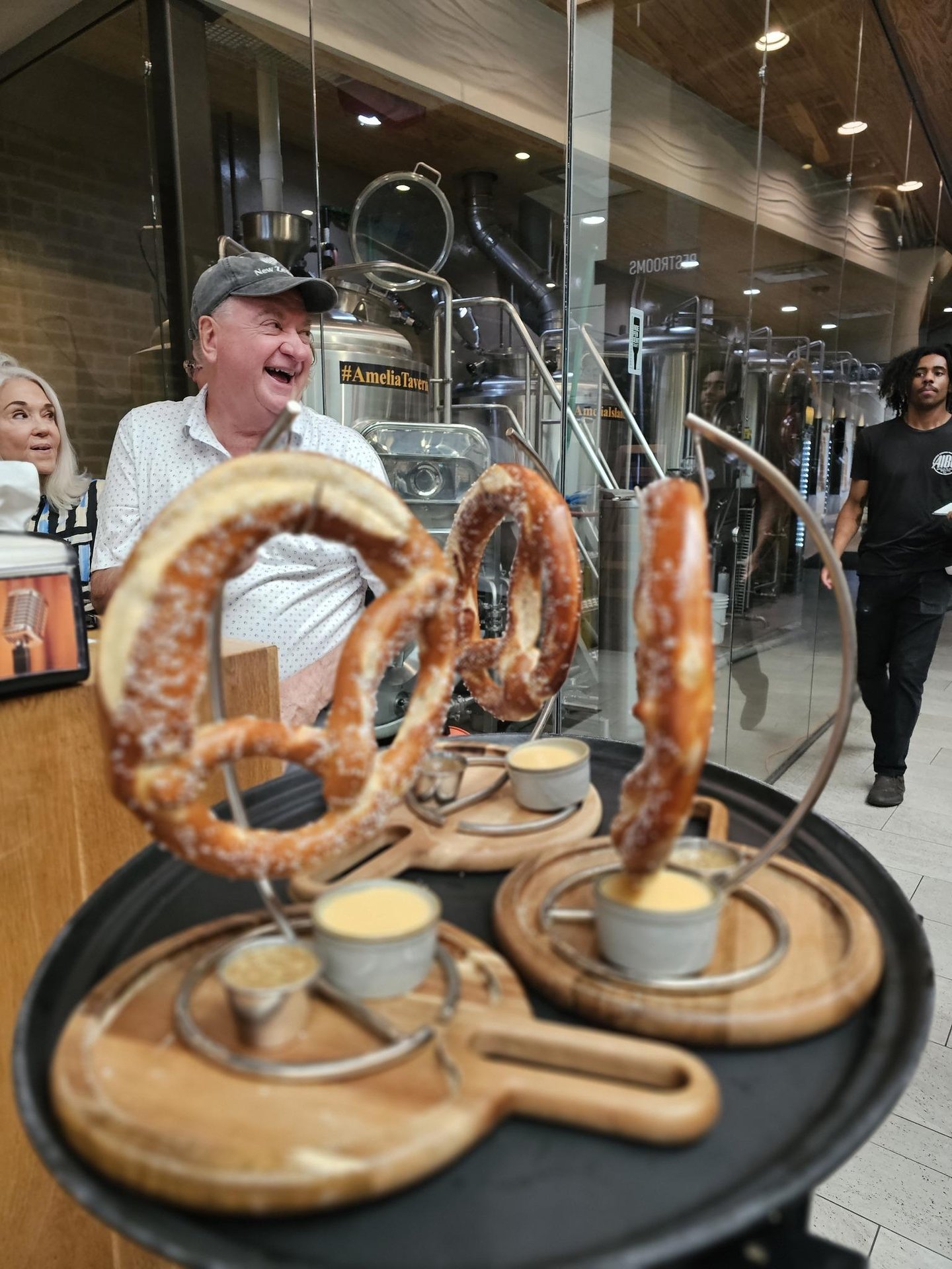 A smiling man stands behind a table holding large pretzels on metal stands, each served with small cups of dipping sauce, in a modern restaurant with glass walls and brewing equipment in the background.