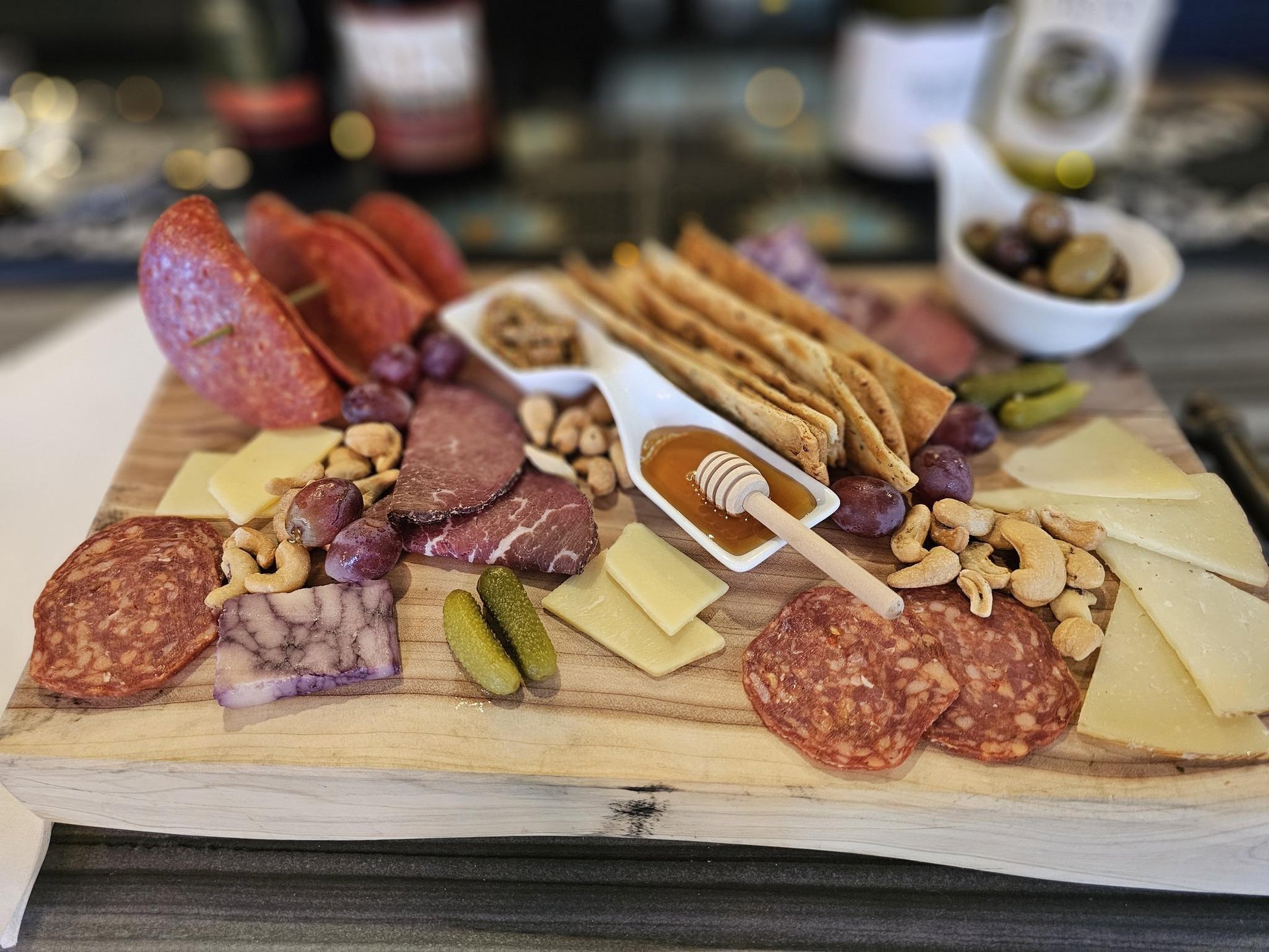A wooden charcuterie board with assorted sliced meats, cheeses, crackers, pickles, grapes, nuts, a small honey dipper with honey, and a bowl of olives, set on a table with wine bottles in the background.