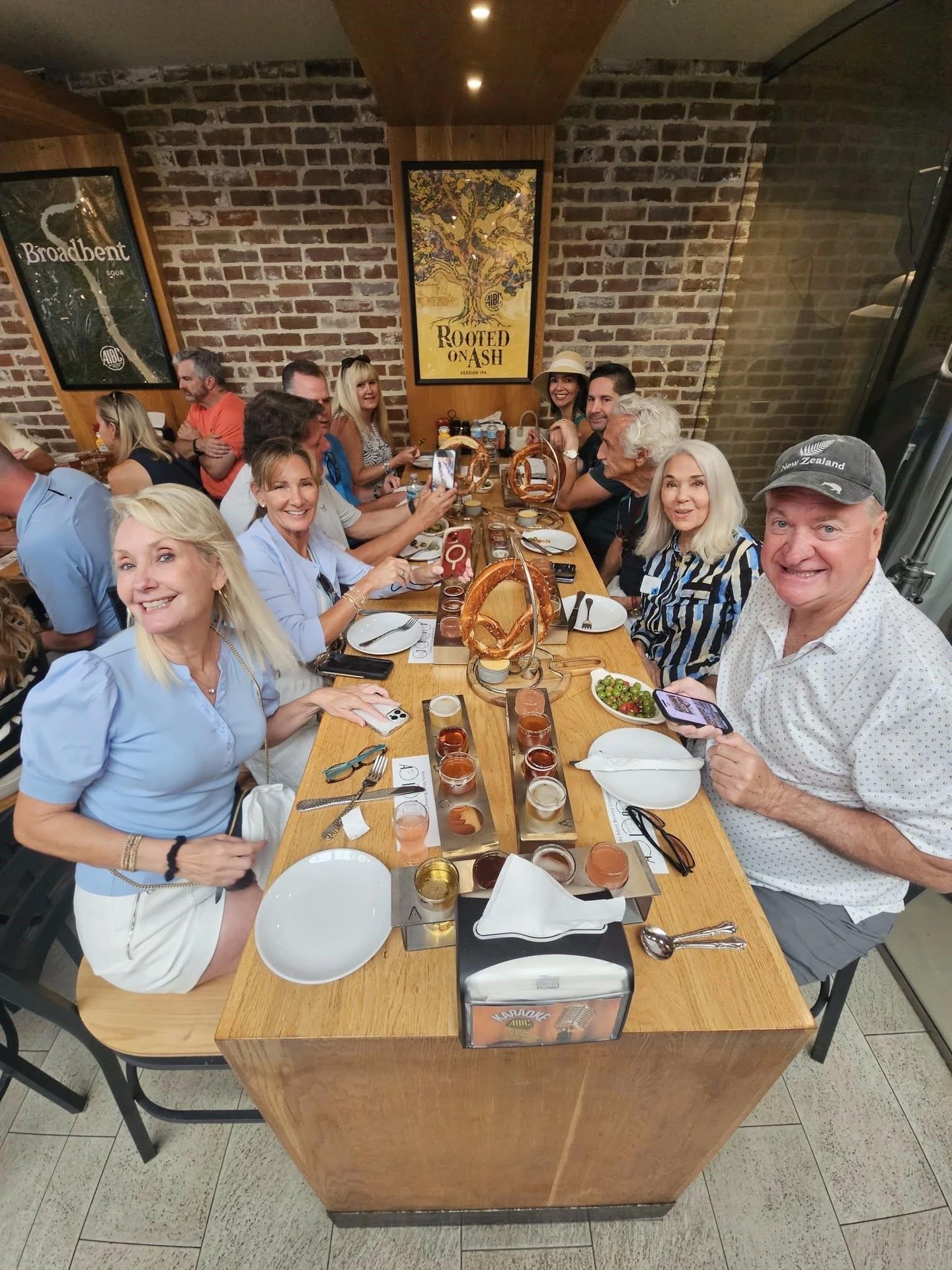 A group of people sit around a long restaurant table, smiling and posing for a photo. The table is set with plates, drinks, and appetizers. The setting features a brick wall and a Rooted in Oak poster in the background.