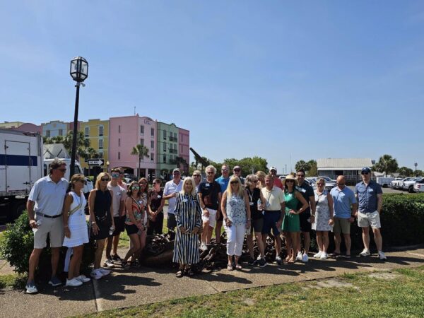 A group of around 25 people stands outdoors on a sunny day, posing for a photo in front of colorful buildings and a tree. Some wear sunglasses and summer clothes. There is a large anchor statue behind them.