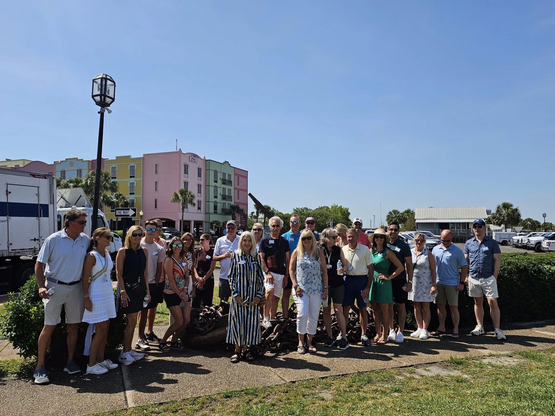 A group of around 25 people stands outdoors on a sunny day, posing for a photo in front of colorful buildings and a tree. Some wear sunglasses and summer clothes. There is a large anchor statue behind them.