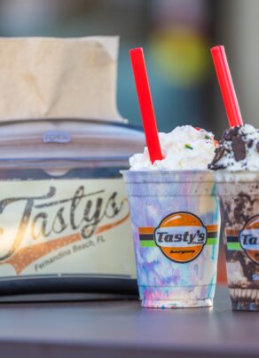 Two milkshakes topped with whipped cream and red straws sit on a table beside a napkin dispenser with the Tasty’s logo. The background is blurred.
