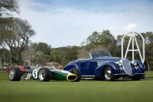 A green and yellow vintage race car with the number 5 is parked next to a blue classic convertible on a grassy field, with trees and a large sculpture in the background.