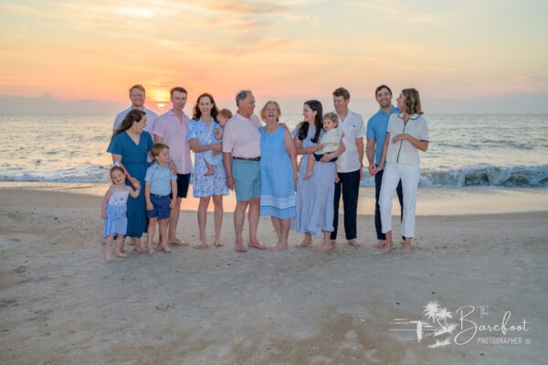 A multigenerational family of twelve, including young children and older adults, stands barefoot on a beach at sunset, smiling and interacting together with the ocean in the background.