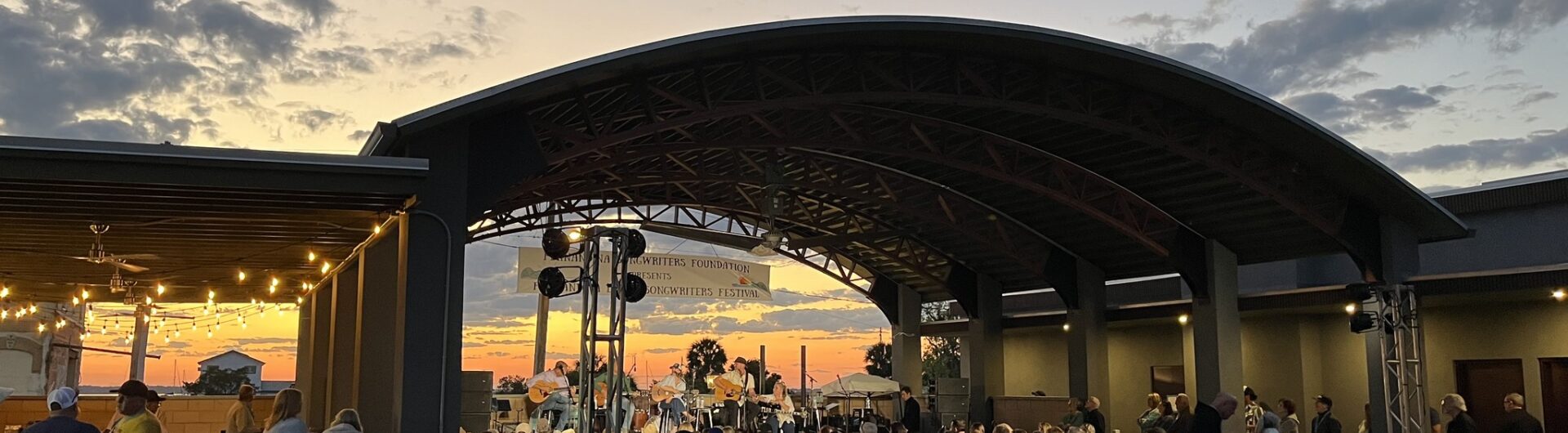 A crowd sits on lawn chairs and blankets, watching a live outdoor concert at sunset under a large, curved pavilion. String lights hang nearby, and the sky is filled with clouds and orange hues.
