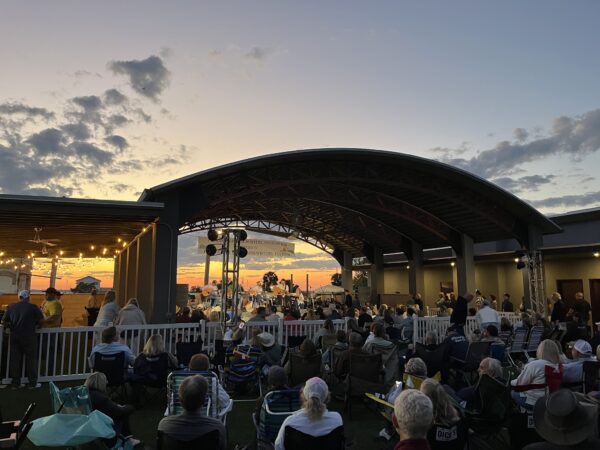 A crowd sits on lawn chairs and blankets, watching a live outdoor concert at sunset under a large, curved pavilion. String lights hang nearby, and the sky is filled with clouds and orange hues.