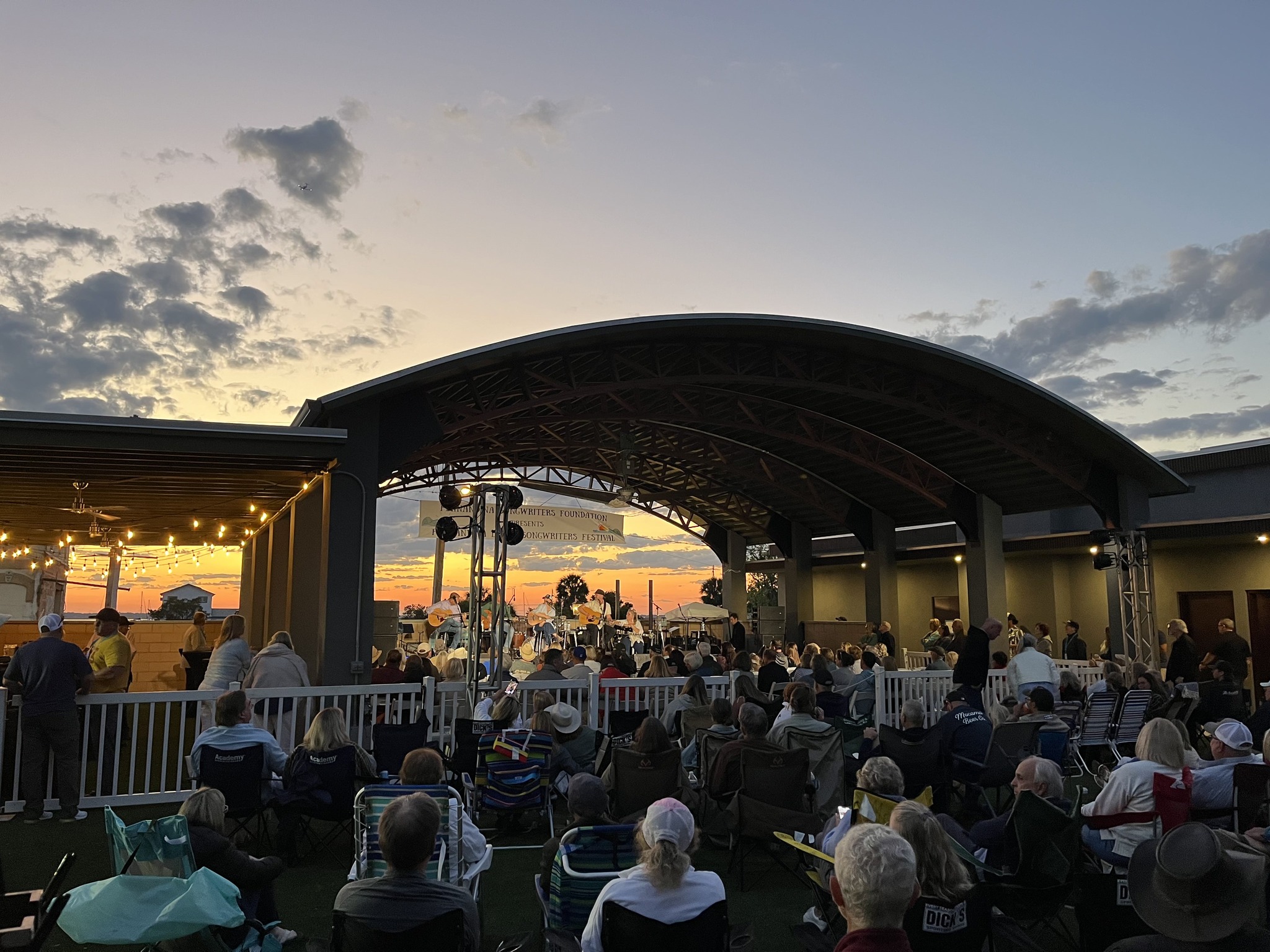 A crowd sits on lawn chairs and blankets, watching a live outdoor concert at sunset under a large, curved pavilion. String lights hang nearby, and the sky is filled with clouds and orange hues.