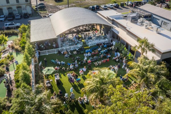 Aerial view of an outdoor event with people sitting on grass and chairs, gathered around a stage with a curved roof. Surrounding areas have palm trees, buildings, and parked cars. The atmosphere is lively and sunny.