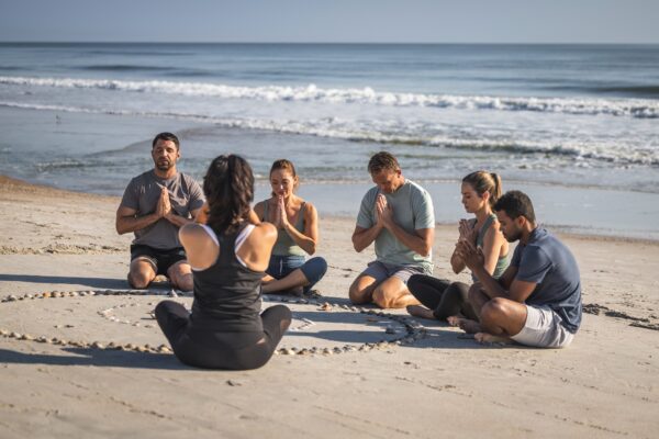 A group of people sit cross-legged in a circle on the beach, eyes closed and hands in prayer position, meditating together near the ocean. A woman faces the group, leading the session.