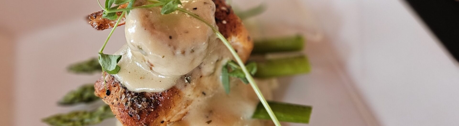 A gourmet dish featuring grilled chicken breast topped with creamy sauce and microgreens, served over mashed potatoes and asparagus spears on a white plate.