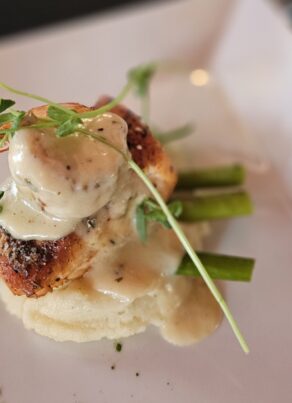 A gourmet dish featuring grilled chicken breast topped with creamy sauce and microgreens, served over mashed potatoes and asparagus spears on a white plate.