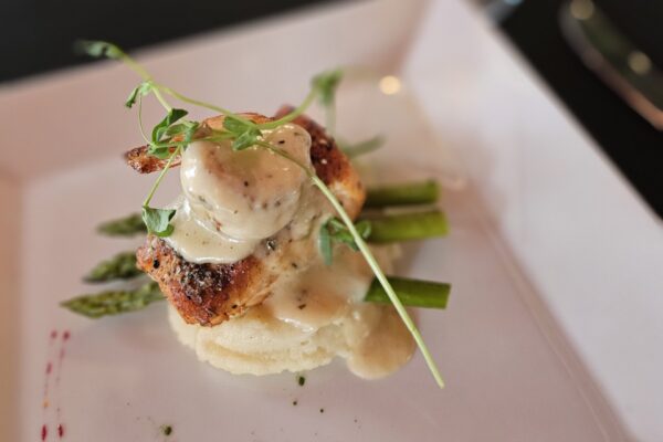 A gourmet dish featuring grilled chicken breast topped with creamy sauce and microgreens, served over mashed potatoes and asparagus spears on a white plate.