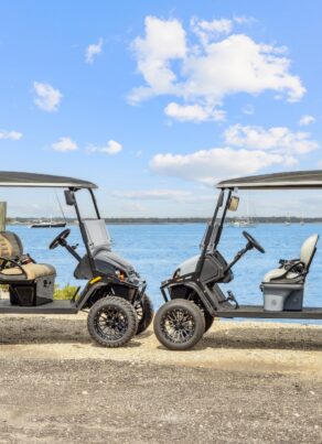 Two black golf carts with beige seats are parked on a gravel area by the water, with a blue sky, scattered clouds, and distant shoreline in the background.