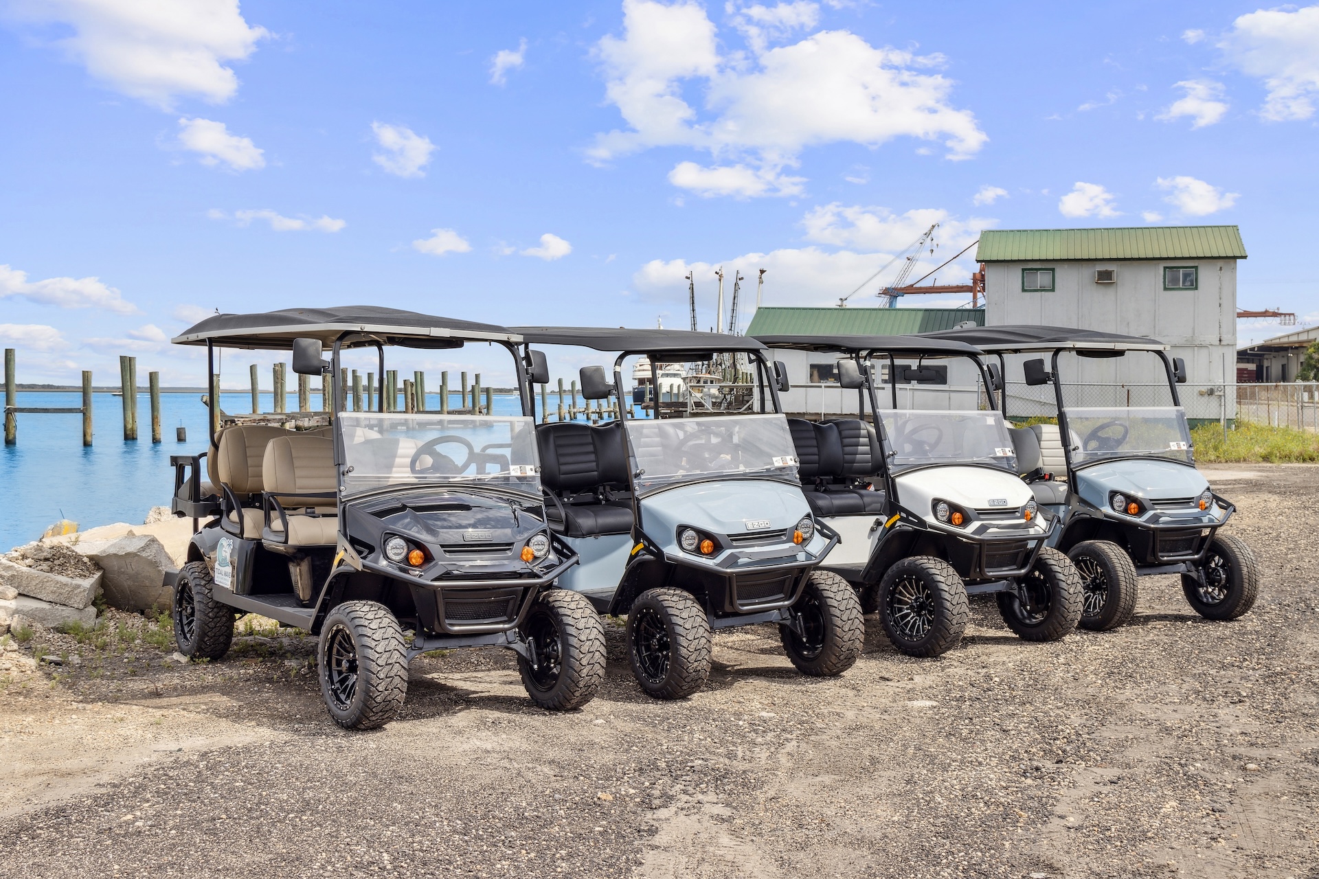 Four golf carts are parked in a row on a gravel lot near a body of water, with docks, wooden posts, and a building with a green roof in the background under a partly cloudy sky.