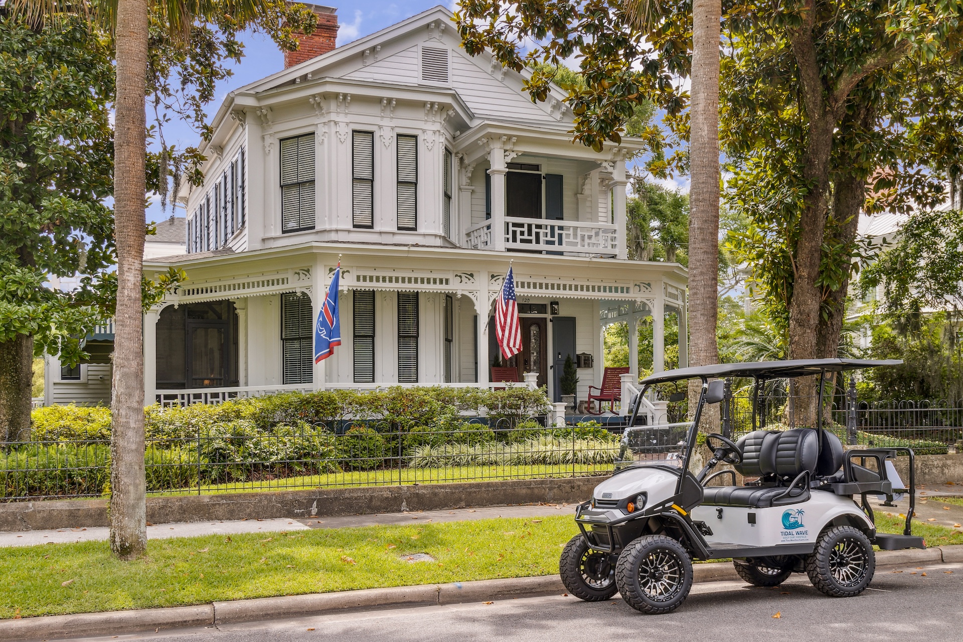 A white, two-story Victorian house with ornate trim, American and state flags, and a porch is surrounded by palm trees. A white golf cart is parked on the street in front of the house.