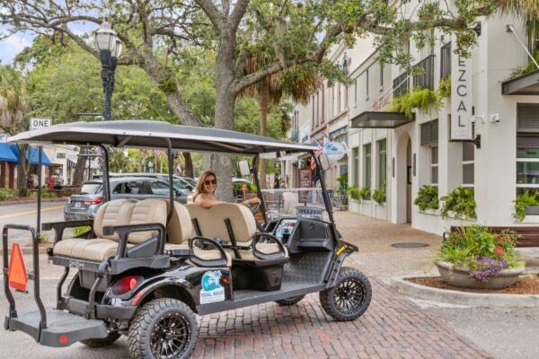 A woman wearing sunglasses sits in a parked golf cart on a sunny, tree-lined street with shops and restaurants, including one named Mezcal, visible in the background.