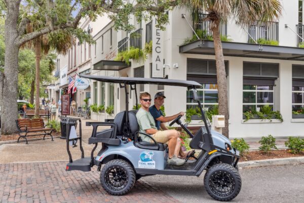 Two men sit in a gray golf cart parked on a brick street in front of shops and restaurants, including one called Mezcal. The area is lined with trees, benches, and potted plants.