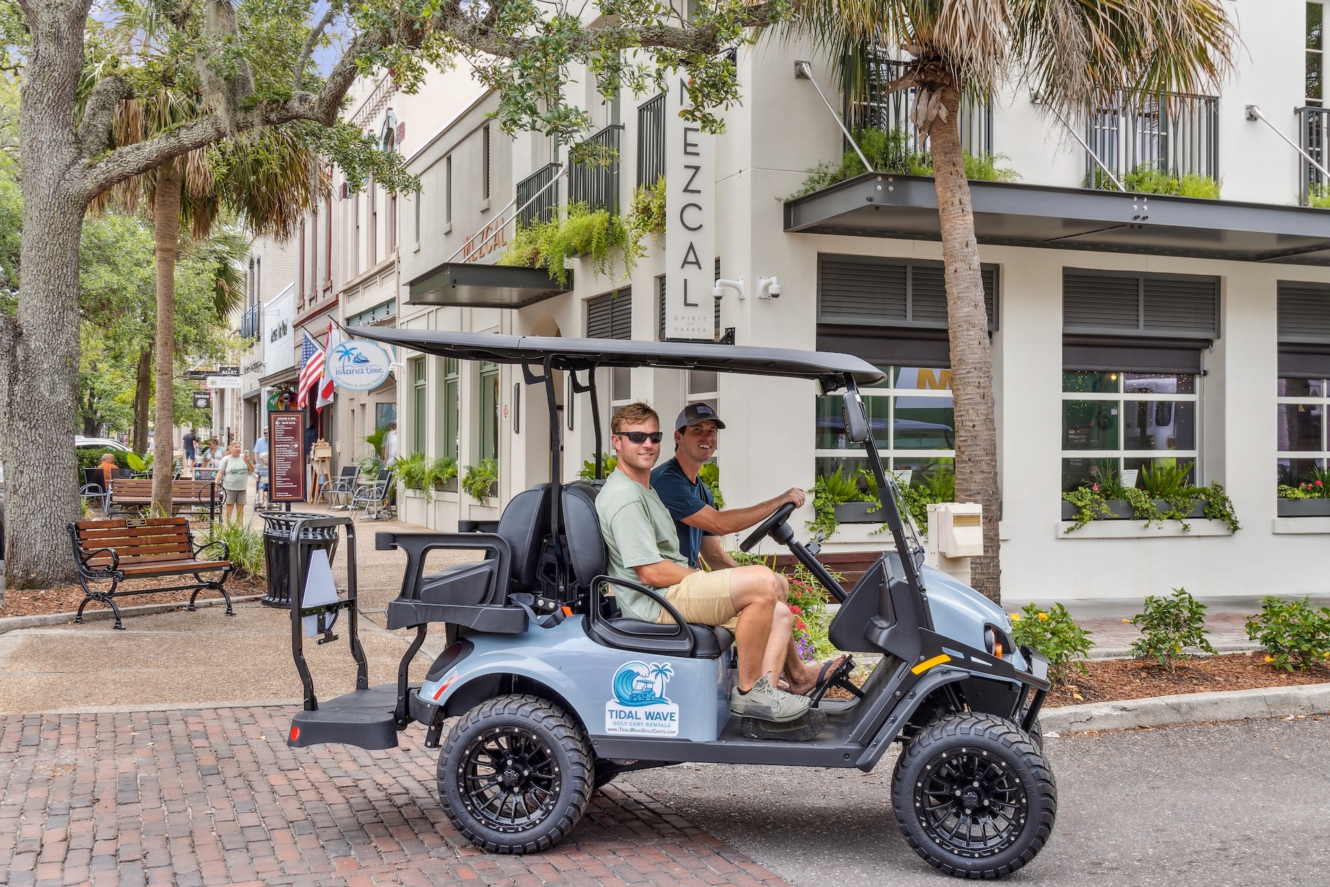 Two men sit in a gray golf cart parked on a brick street in front of shops and restaurants, including one called Mezcal. The area is lined with trees, benches, and potted plants.