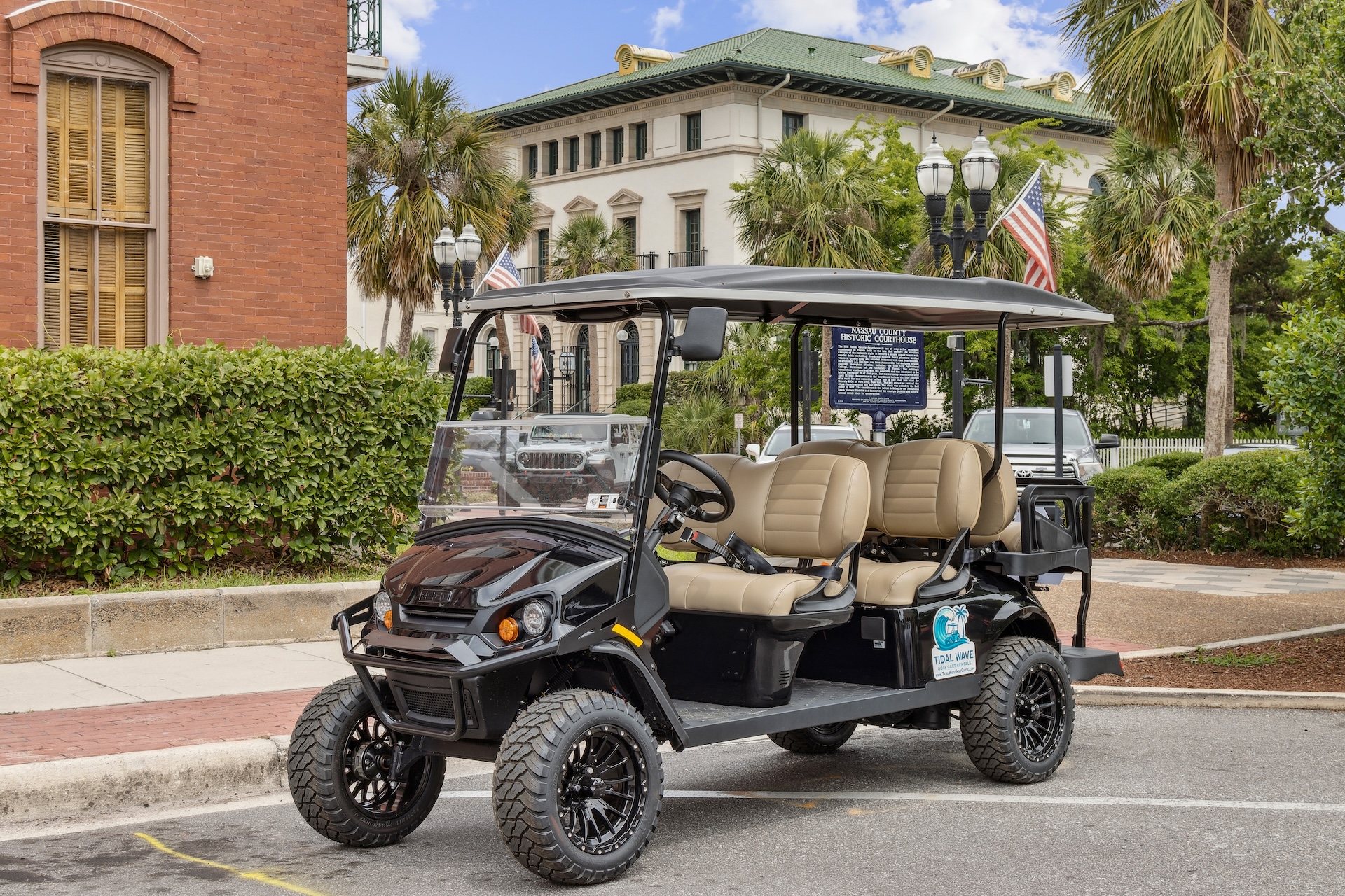 A black golf cart with beige seats is parked on a city street, surrounded by greenery and historic buildings. Two American flags are visible on the cart’s back, and palm trees are in the background.