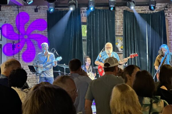 A band performs on stage in front of a crowd, with blue and purple lights shining. The musicians play guitars and drums, and a pink flower design is projected on a brick wall behind them. Audience members watch and enjoy the music.