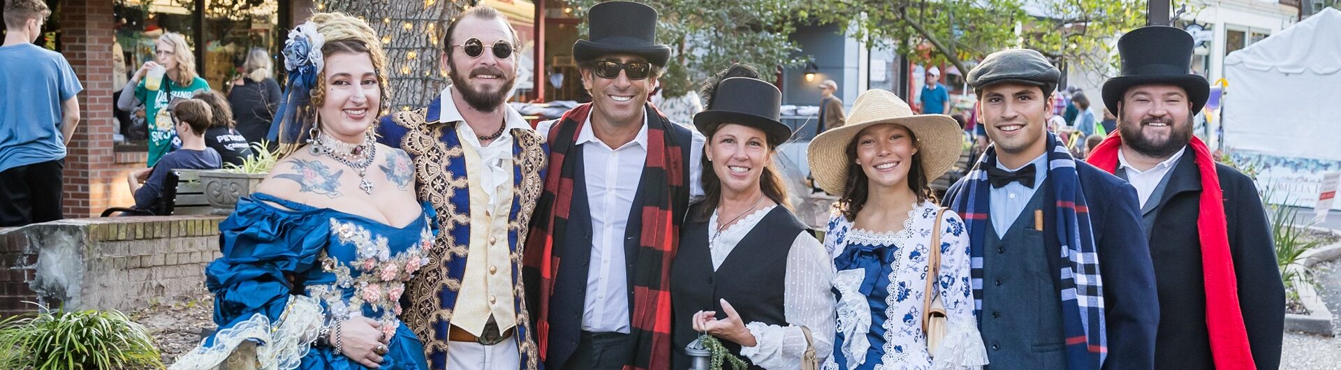 A group of seven people in historical and Victorian-style costumes stand together outdoors on a street, smiling at the camera. Trees, brick buildings, and a streetlamp are visible in the background.