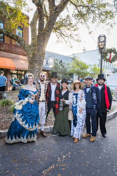 A group of seven people in historical and Victorian-style costumes stand together outdoors on a street, smiling at the camera. Trees, brick buildings, and a streetlamp are visible in the background.