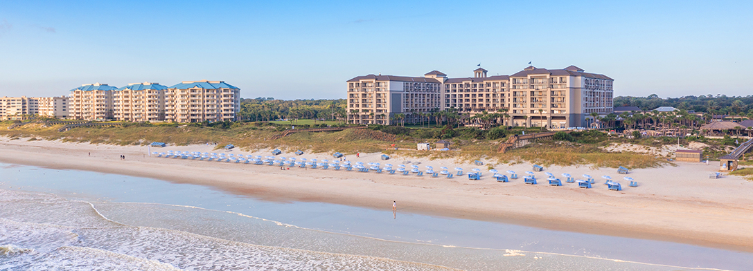 A wide sandy beach lined with rows of white beach chairs and umbrellas, in front of large beachfront hotels and condos, with greenery and a clear blue sky in the background.