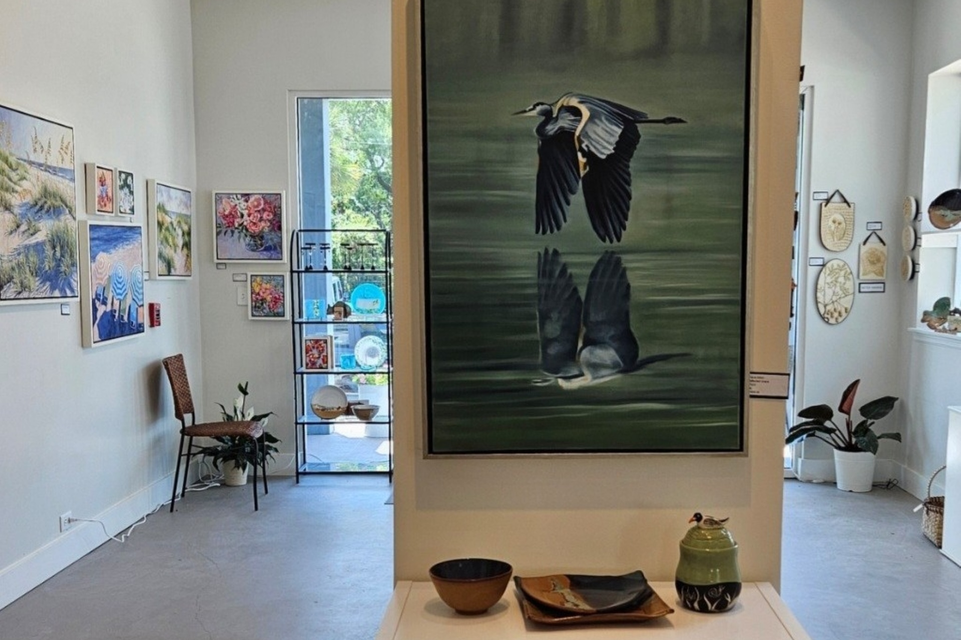 A bright art gallery displays various colorful paintings and ceramics. In the center, a large painting of a bird flying over water with its reflection is showcased. Potted plants and chairs are arranged around the space.