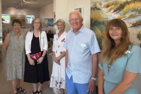 Five adults stand smiling inside an art gallery, with paintings on the walls and natural light coming through the windows. They are dressed in casual and semi-formal summer attire.