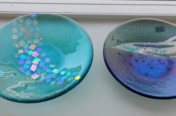 Two round, decorative glass dishes with abstract designs. The left dish is teal with iridescent mosaic squares, and the right dish is blue and purple with a textured, wave-like pattern. Both are displayed on a white surface.