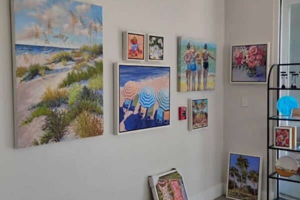 A corner of a room displays colorful beach-themed and floral paintings on the walls, along with a few framed artworks resting on the floor and a black metal shelf with decorative items nearby.