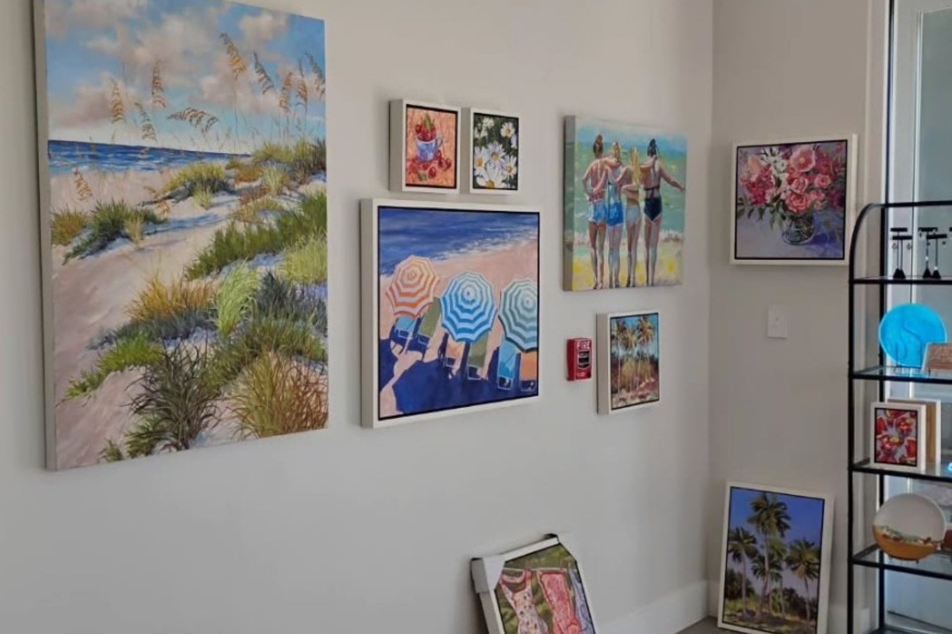 A corner of a room displays colorful beach-themed and floral paintings on the walls, along with a few framed artworks resting on the floor and a black metal shelf with decorative items nearby.