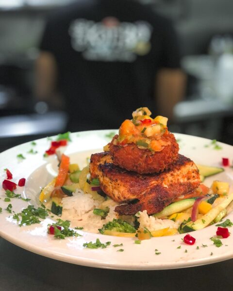 A white plate with grilled fish and a golden-fried seafood cake on top, served over white rice and sautéed vegetables, garnished with herbs and diced peppers. A blurred person stands in the background.