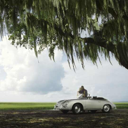 A person sits in a vintage white convertible parked under a large tree with hanging Spanish moss, overlooking a serene landscape of green fields and water under a bright sky with scattered clouds.