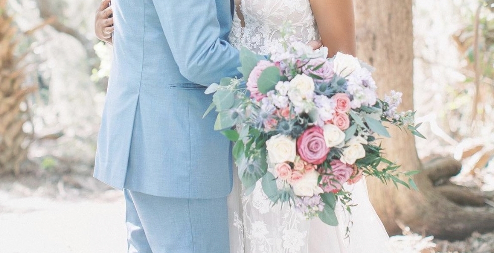 A couple dressed in wedding attire stands close together outdoors. The bride holds a large bouquet of pink, purple, and white flowers with greenery. The groom wears a light blue suit. Trees and sunlight fill the background.