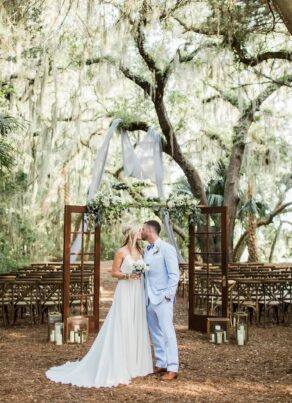 A bride and groom stand kissing in front of a rustic outdoor wedding altar, surrounded by trees draped with Spanish moss, wooden chairs, and lanterns, under a canopy of greenery and white fabric.