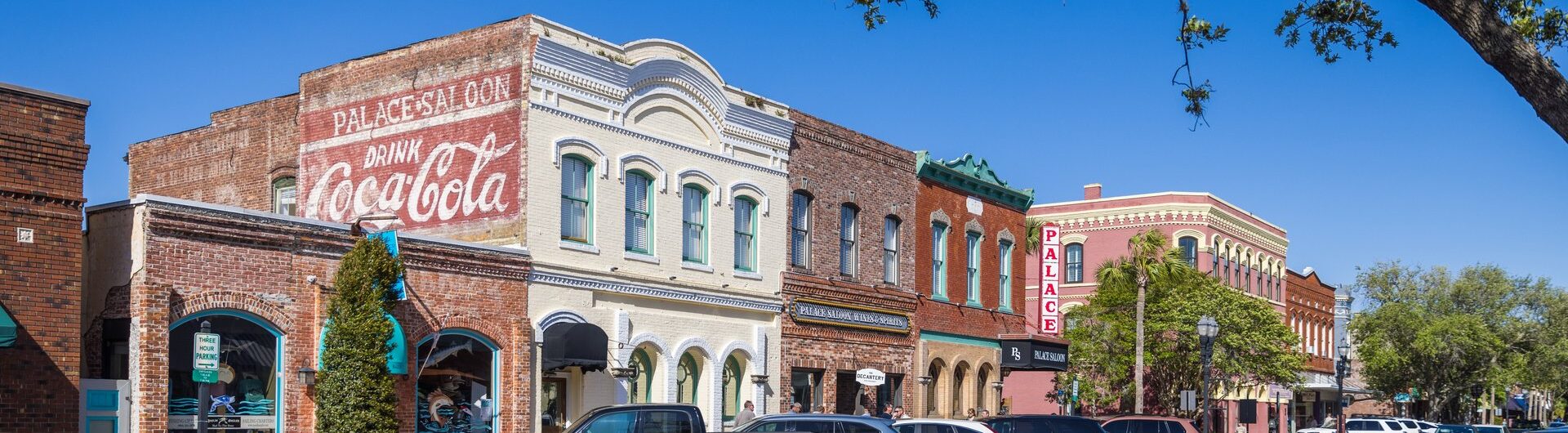A row of historic brick buildings with colorful facades and vintage Coca-Cola signage line a sunny small-town street, with parked cars and trees framing the scene under a clear blue sky.