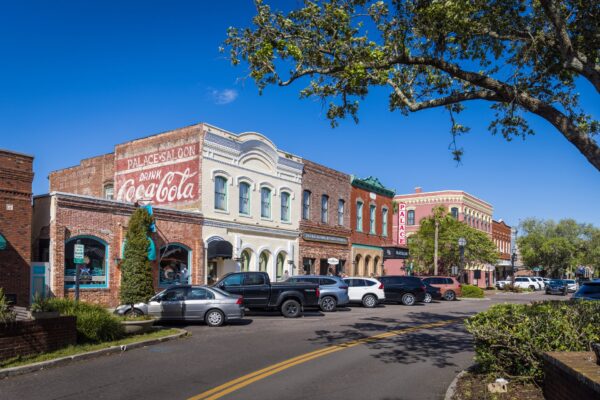 A row of historic brick buildings with colorful facades and vintage Coca-Cola signage line a sunny small-town street, with parked cars and trees framing the scene under a clear blue sky.
