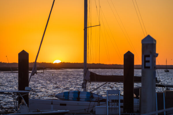 A vibrant orange sunset over a calm marina with silhouetted sailboats and dock posts in the foreground. The sun is partially visible on the horizon, casting a warm glow on the water.