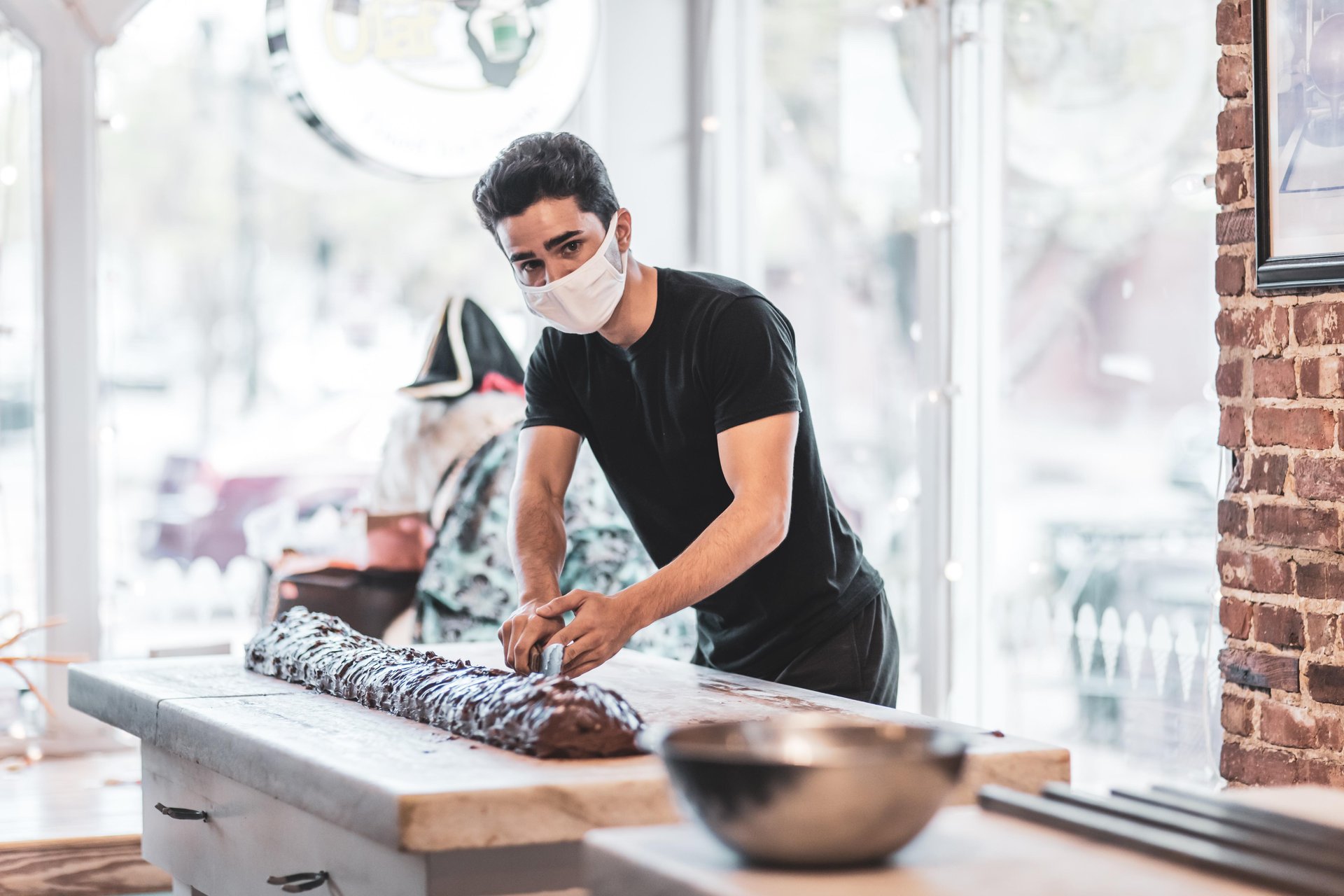 A man wearing a face mask and black shirt shapes a long piece of chocolate on a marble countertop in a bright, modern kitchen, with a large metal bowl nearby.