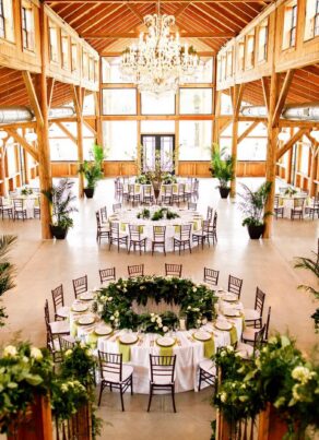A spacious, rustic banquet hall with wooden beams, large windows, round tables set for an event, greenery decorations, white tablecloths, brown chairs, potted plants, and a crystal chandelier overhead.